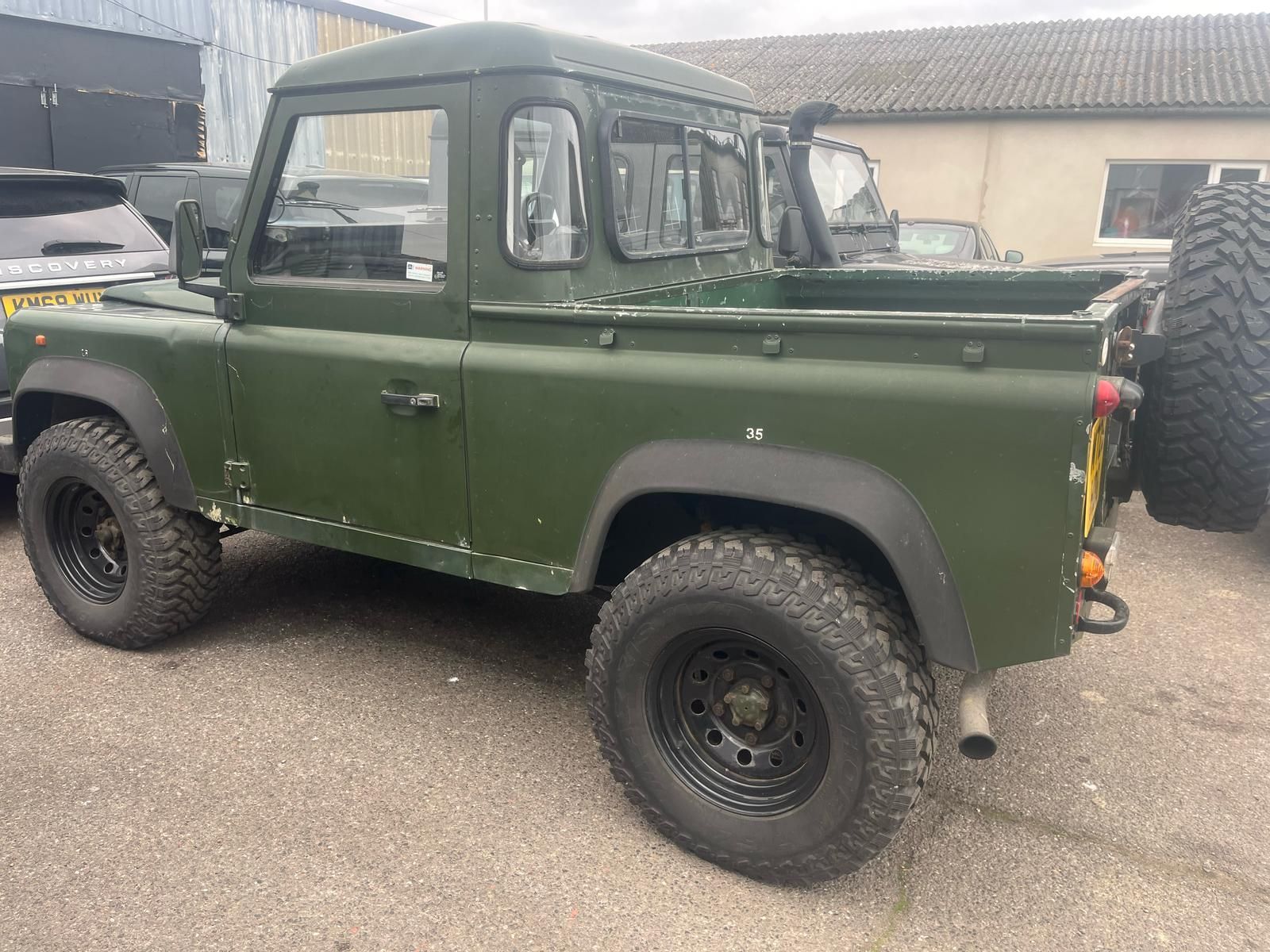 Green Land Rover Defender pickup truck parked outdoors on pavement. It has black wheels and a roll bar.