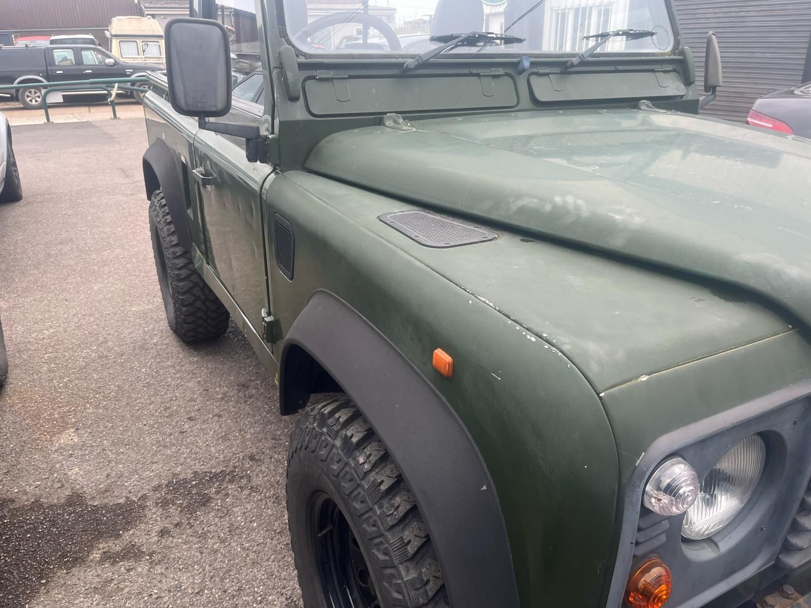 Green Land Rover Defender parked on a paved surface. Shows front-left side with black fender flares and tires.