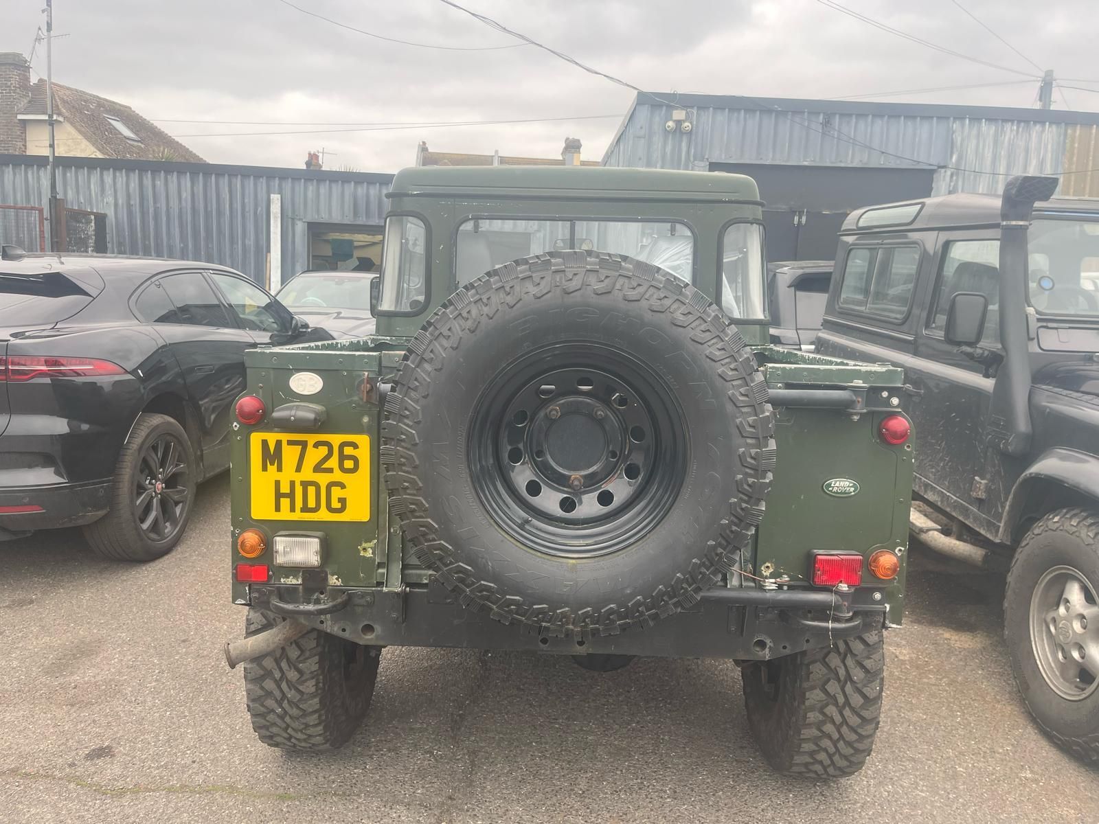 Rear view of a green Land Rover Defender with a spare tire, parked outdoors, license plate reads M726 HDG.