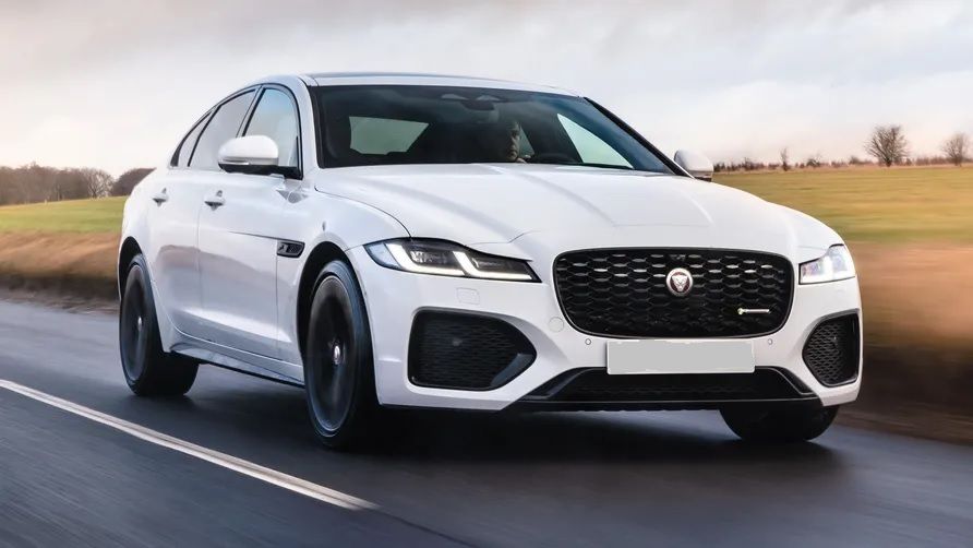 White Jaguar XF sedan driving on a road, front view, black grill and wheels, daytime.