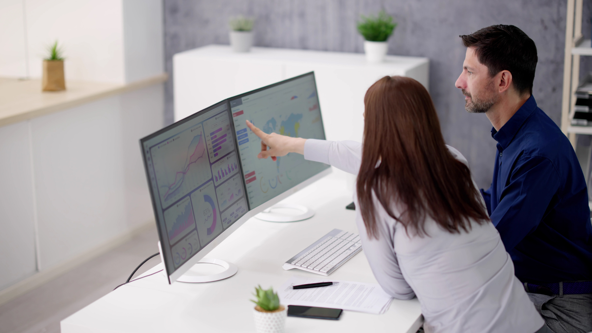 A man and a woman are sitting at an office desk, looking at a screen with data analytics on.