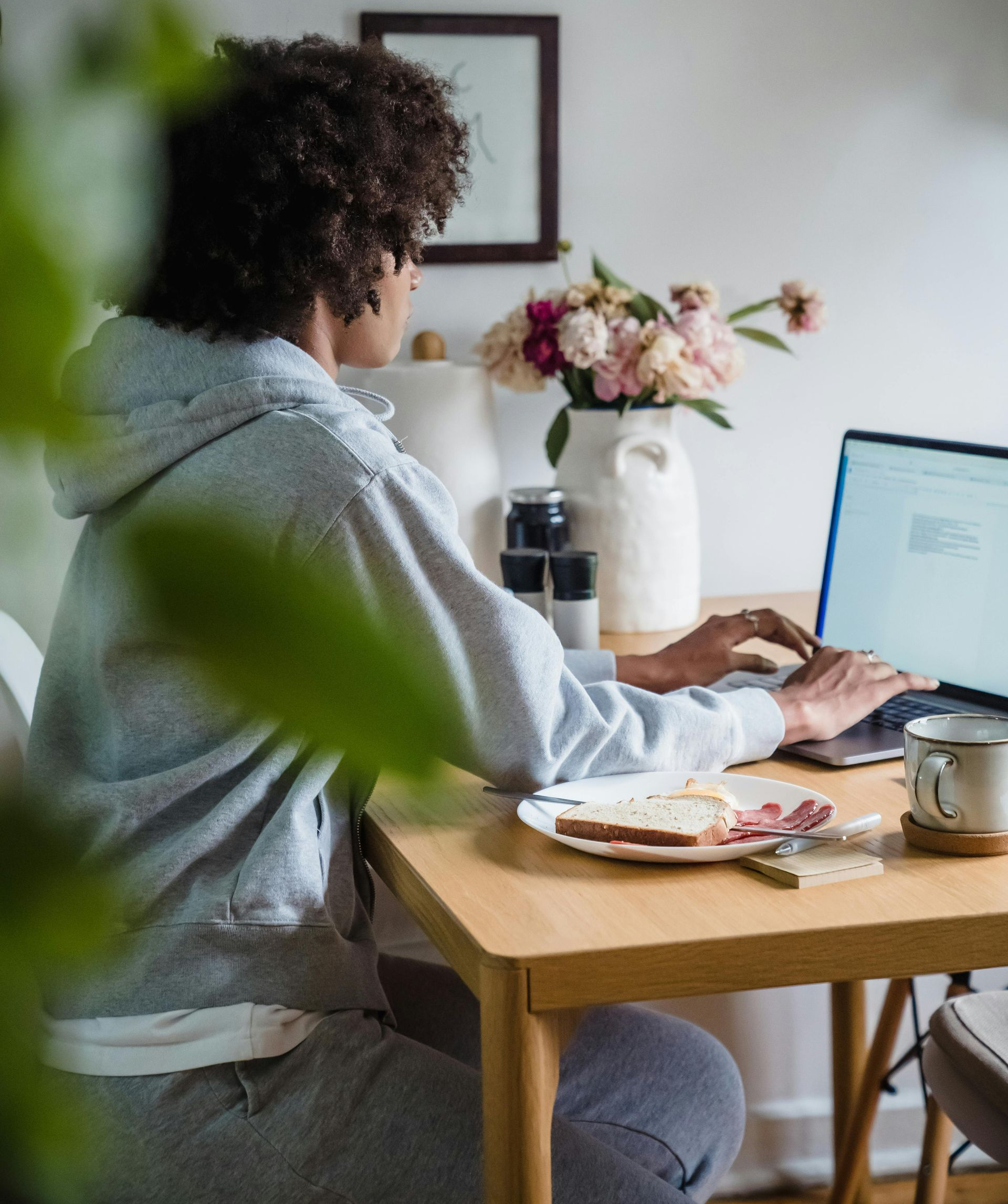 Person in a gray hoodie works on a laptop at a table, breakfast plate and coffee nearby.