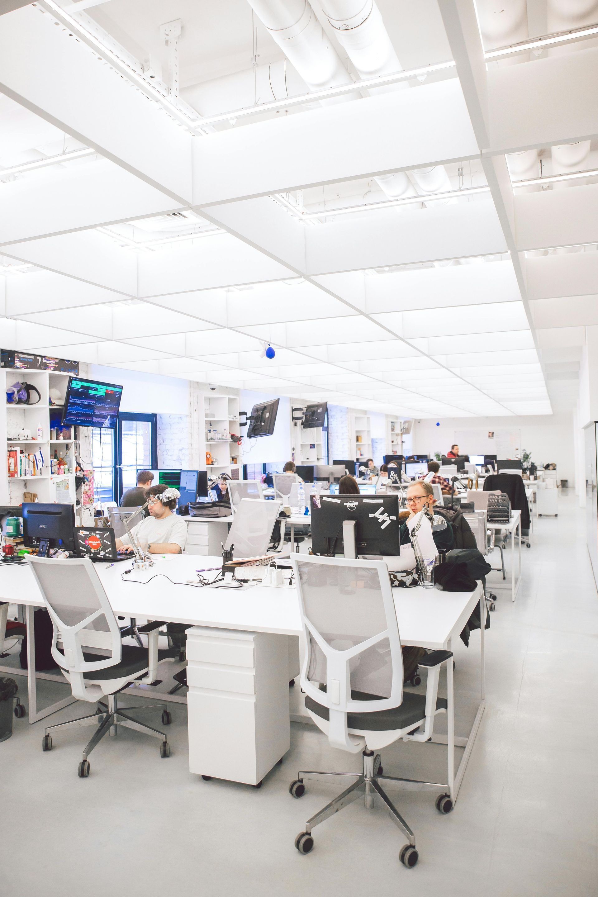 Bright, modern office space with white desks and chairs. People work at computers under a grid ceiling.