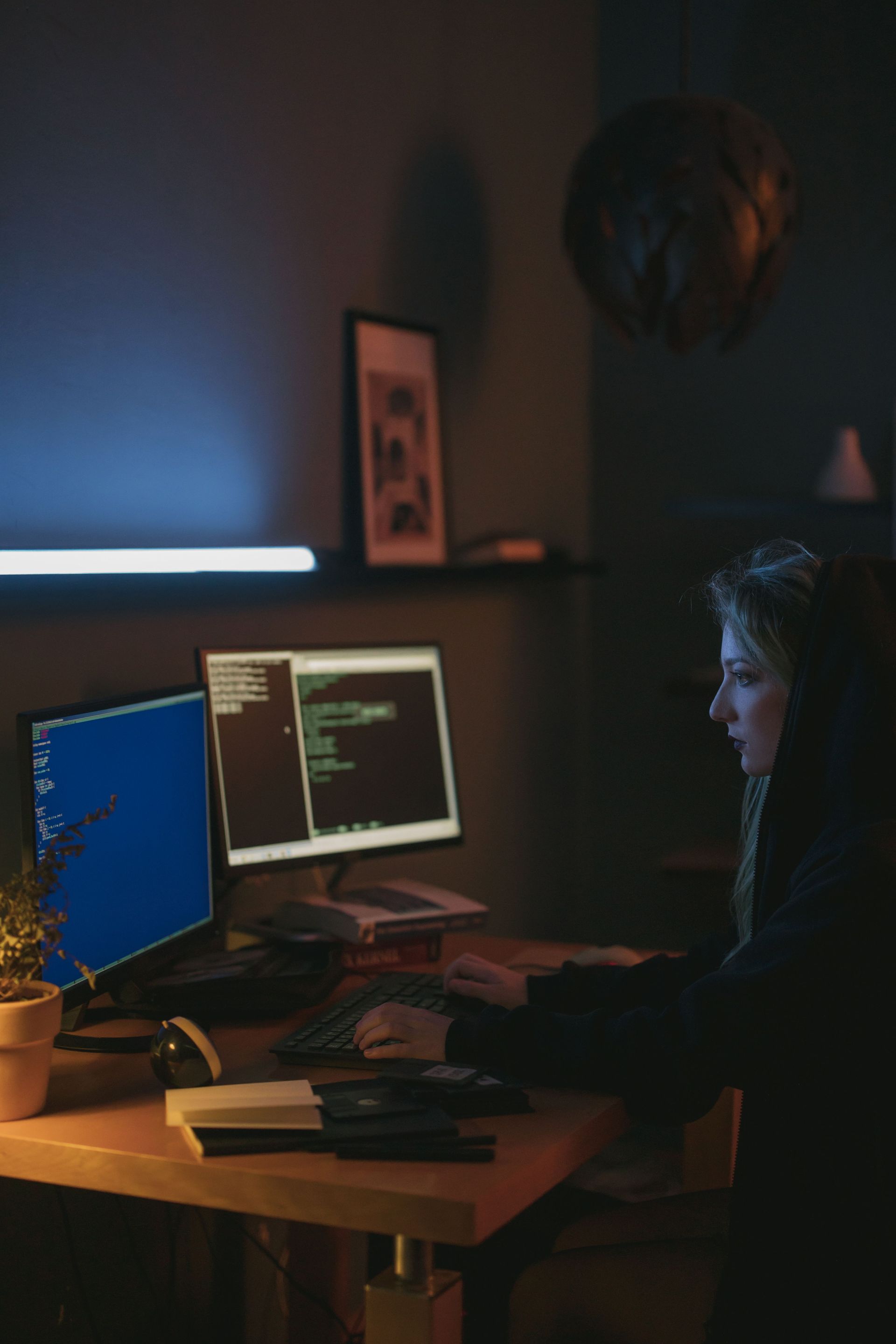 Person working at a desk with dual monitors in a dimly lit room, lit by blue screen glow.