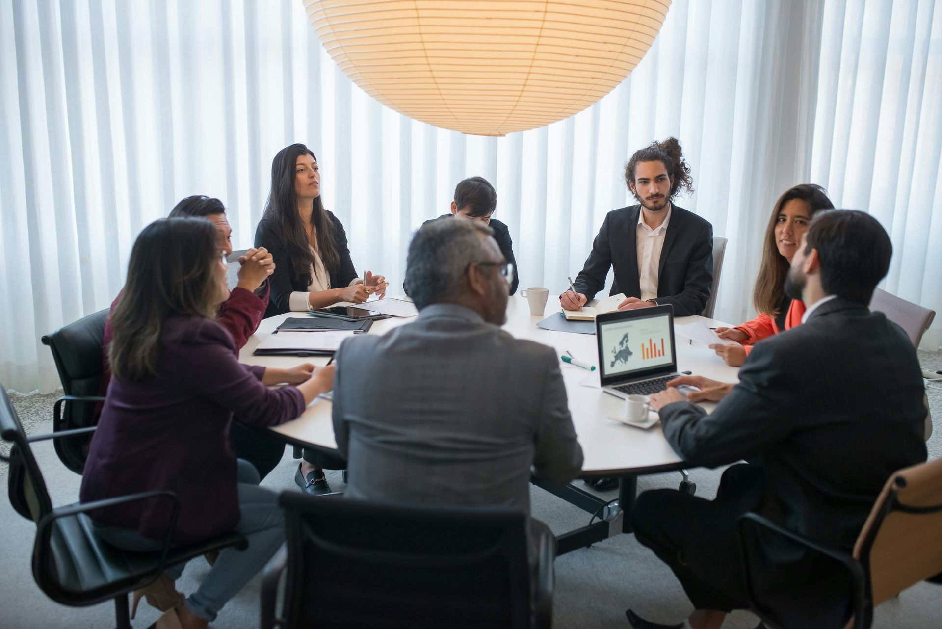 People in business attire seated around a table, laptop with a graph visible, in a modern office setting.