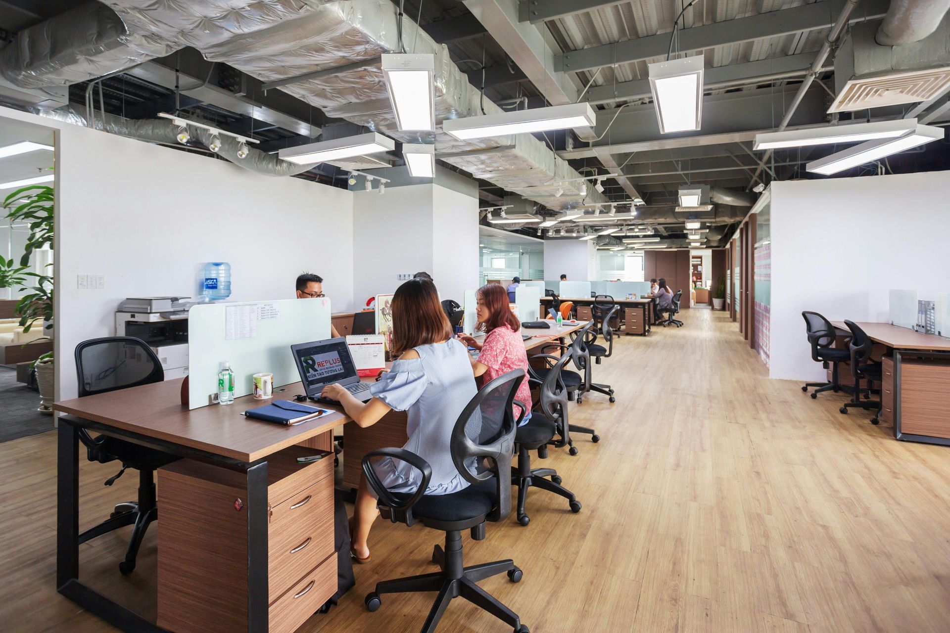 Open-plan office with people working at desks, wood flooring, and exposed ceiling beams