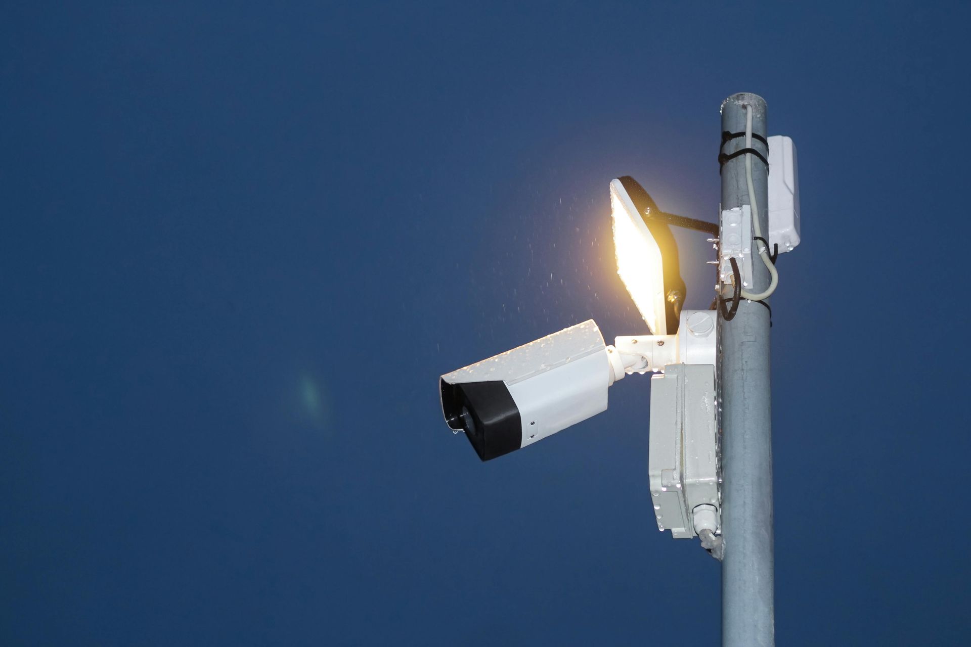 Security camera and light fixture mounted on a metal pole against a dark blue sky.