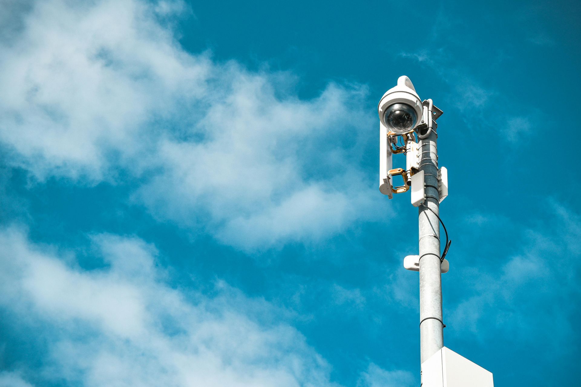 A security camera and communication antennas mounted on a white pole against a bright blue cloudy sky.