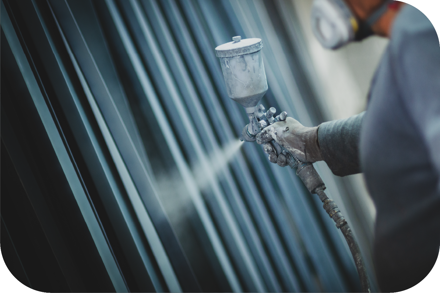 A worker wearing a respirator uses a spray gun to apply a finish to dark metal panels.