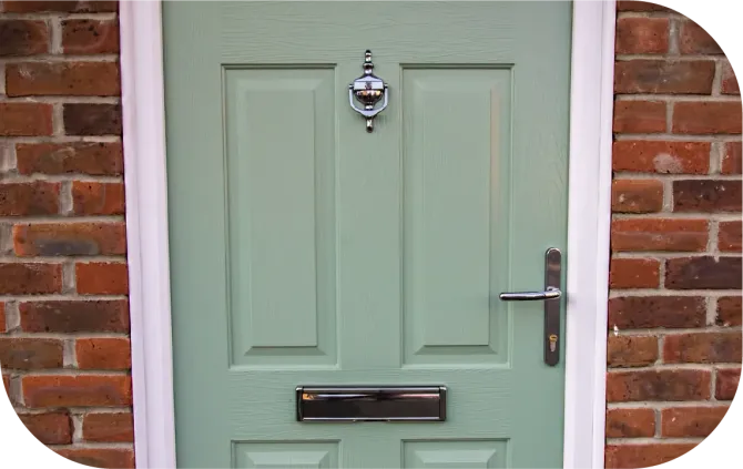 A sage green front door with a silver knocker, a black letterbox, and a handle, set within a red brick wall.