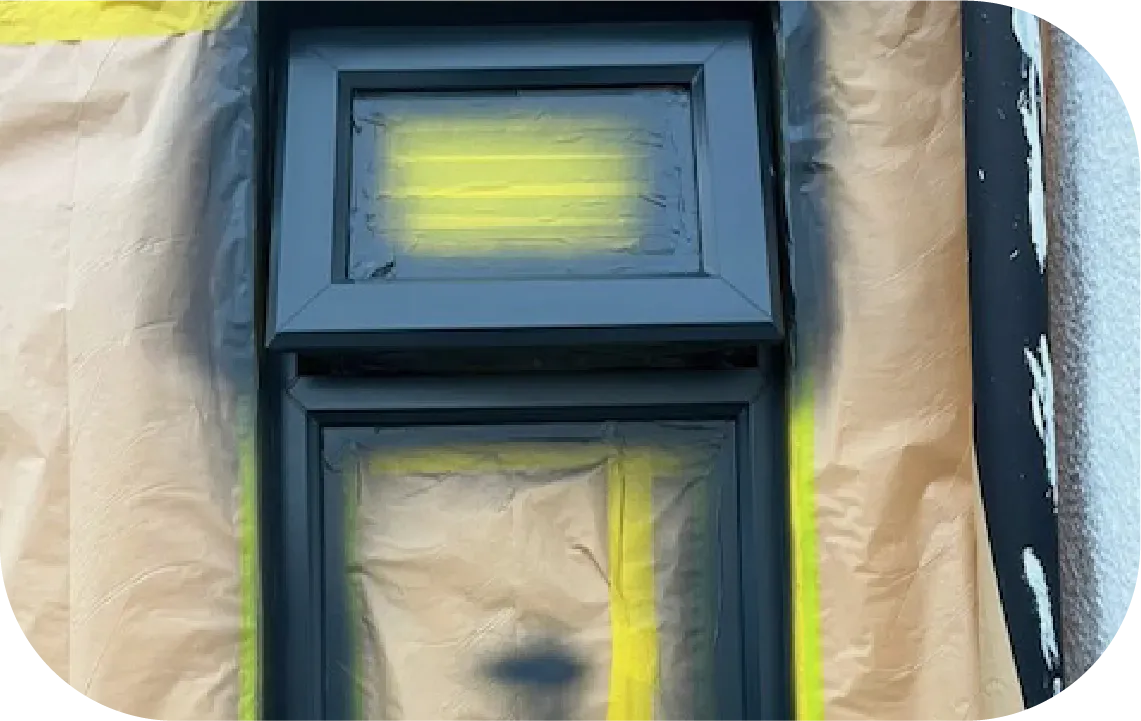 A black window frame being spray-painted, surrounded by yellow masking tape and brown protective paper.