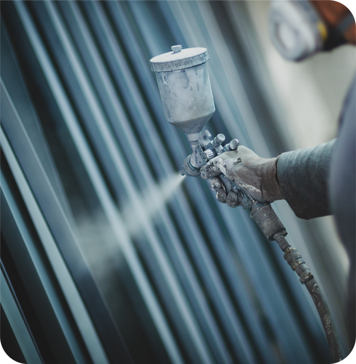 A person wearing a protective mask uses a spray gun to apply a gray coating to a stack of metal beams.