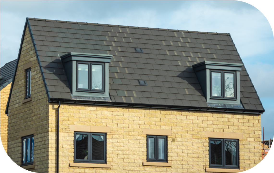 A tan stone house with a dark gray shingled roof, featuring two dormer windows and several smaller windows.