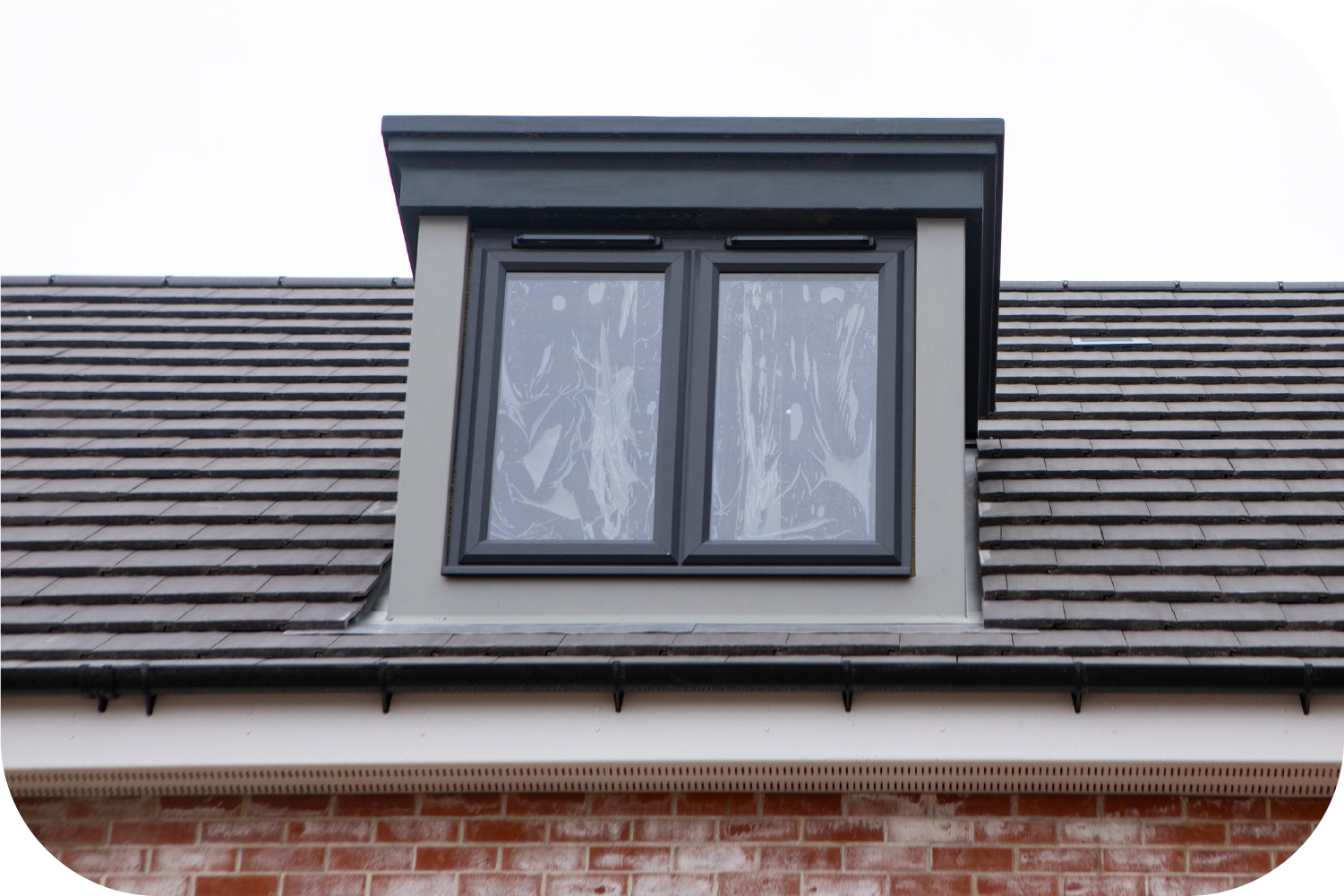 A dormer window with a dark frame set into a gray tiled roof above a brick building wall.