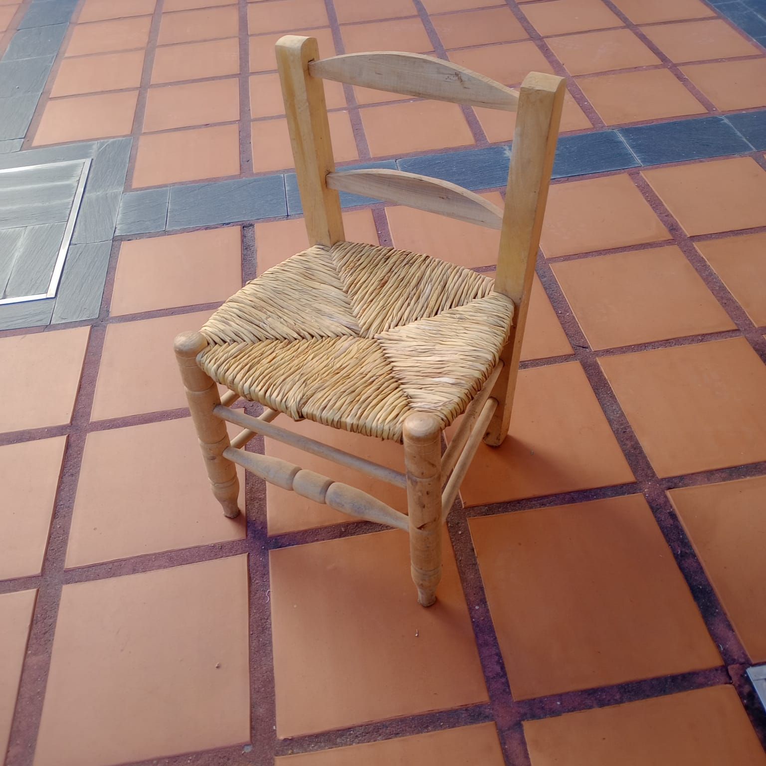 Una pequeña silla de madera con asiento de junco tejido se encuentra sobre un suelo de baldosas de terracota.