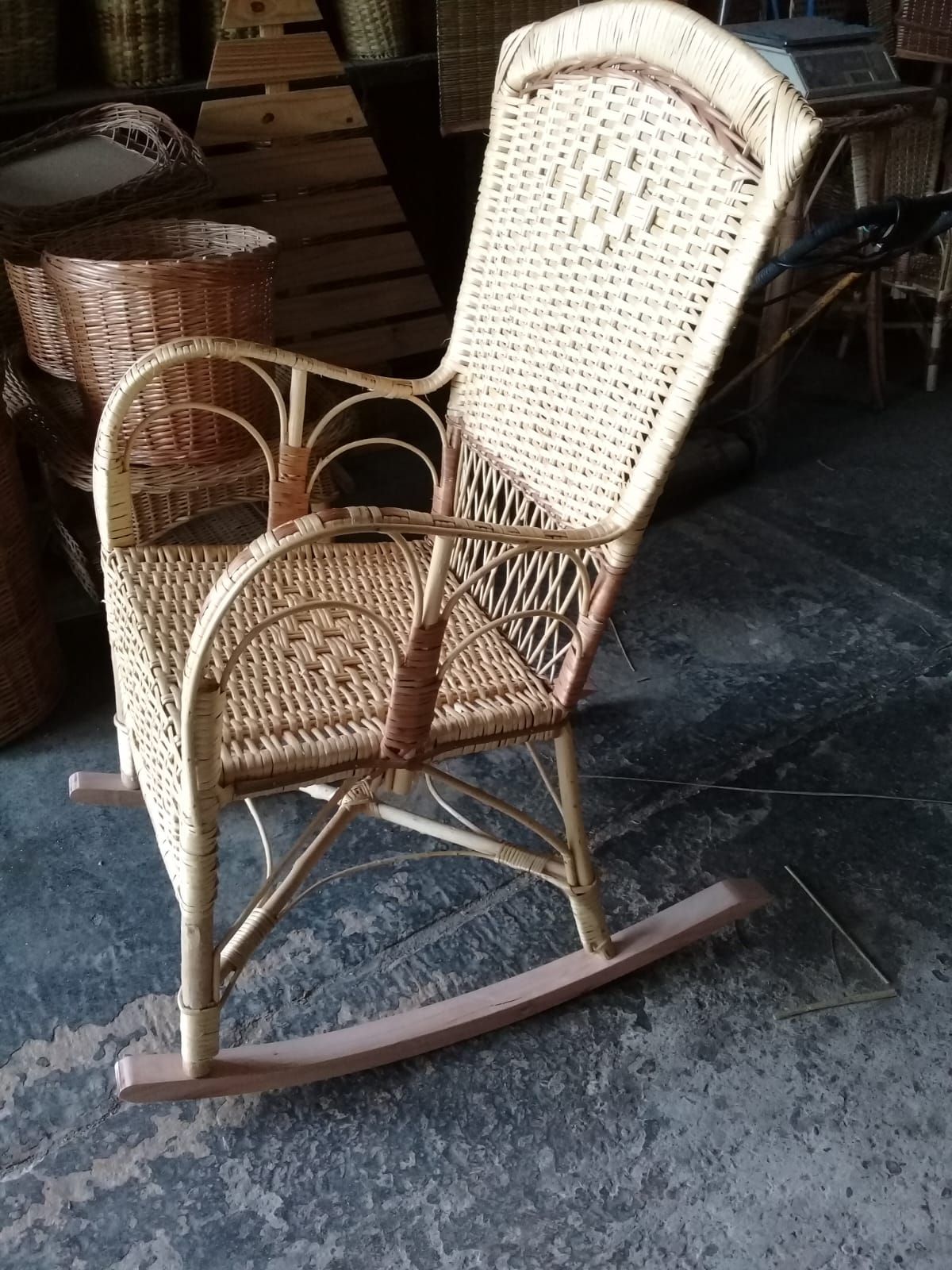 Una mecedora de mimbre de color claro se alza sobre un suelo de hormigón gris, con cestas tejidas al fondo.