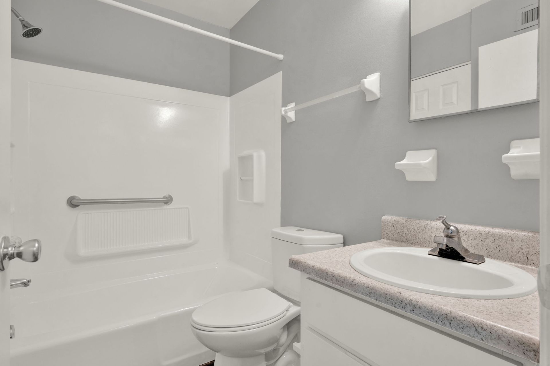Bathroom with a white tub/shower, toilet, sink on a speckled countertop, and gray walls.