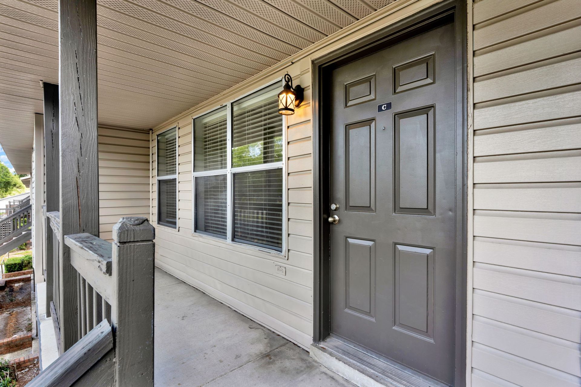 Exterior view of a beige building with a gray door, windows, and a porch with a wooden railing.