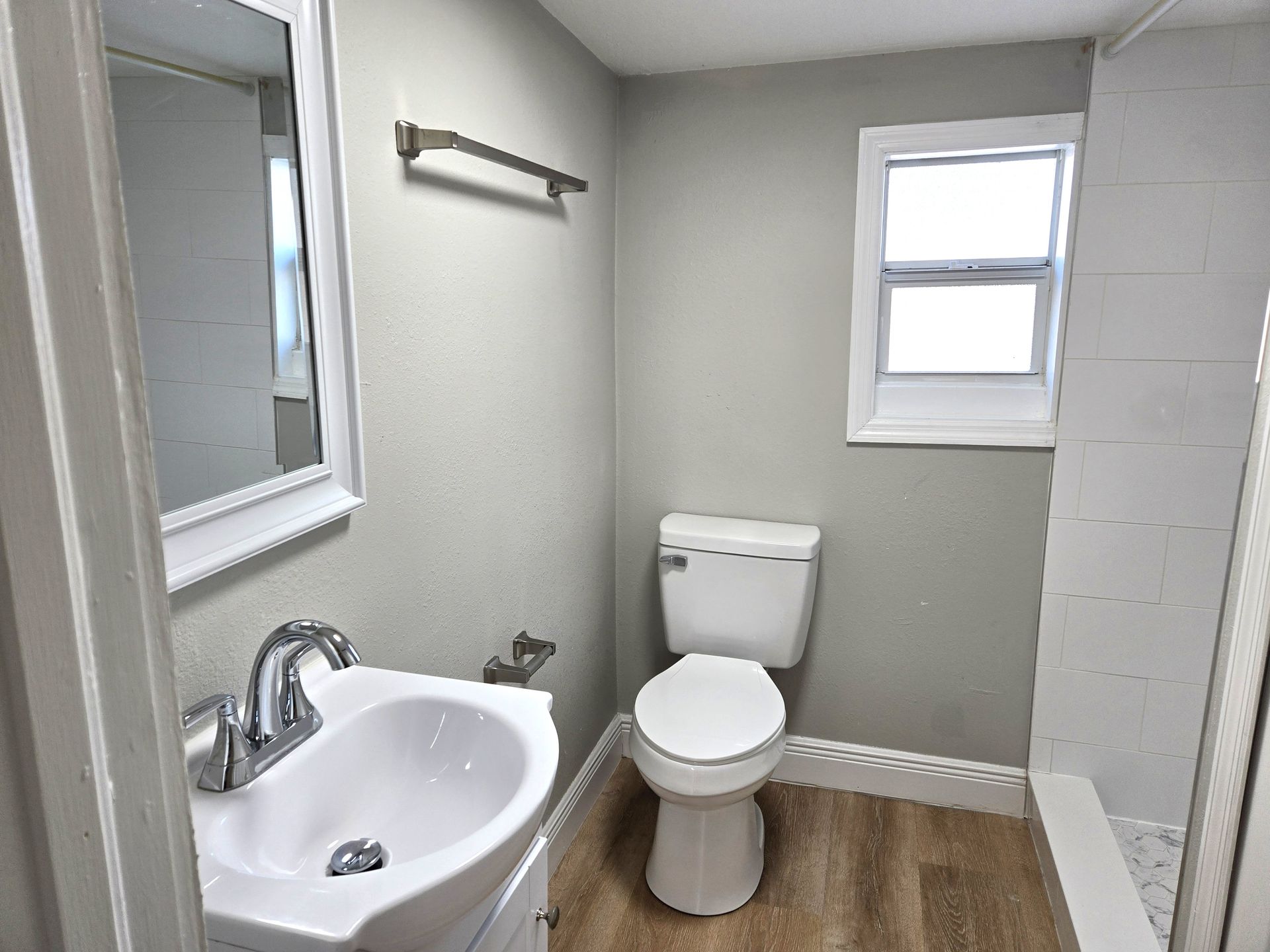 Small bathroom with toilet, sink, and window; light gray walls and wood-look flooring.