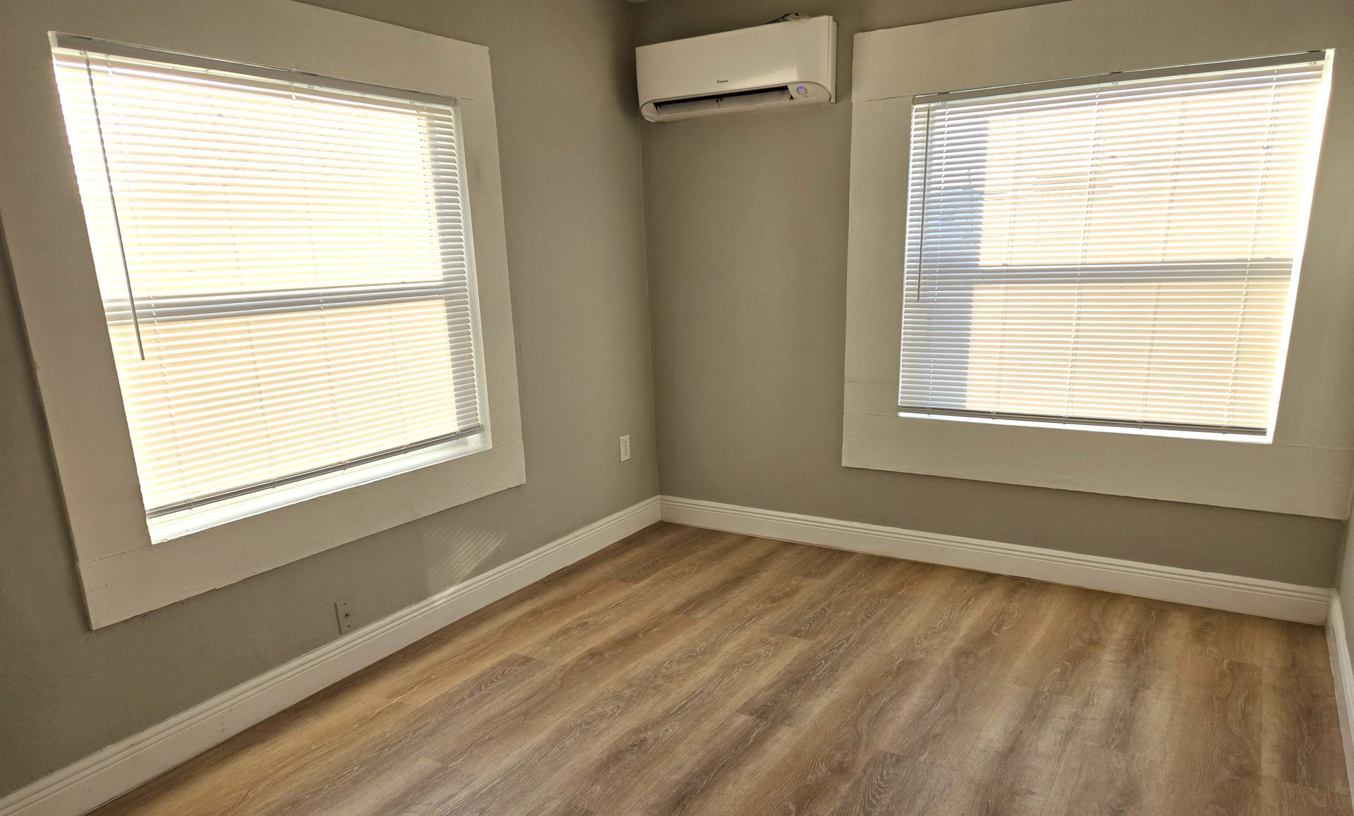 Empty room with two windows, light gray walls, wood-look floor, air conditioner.