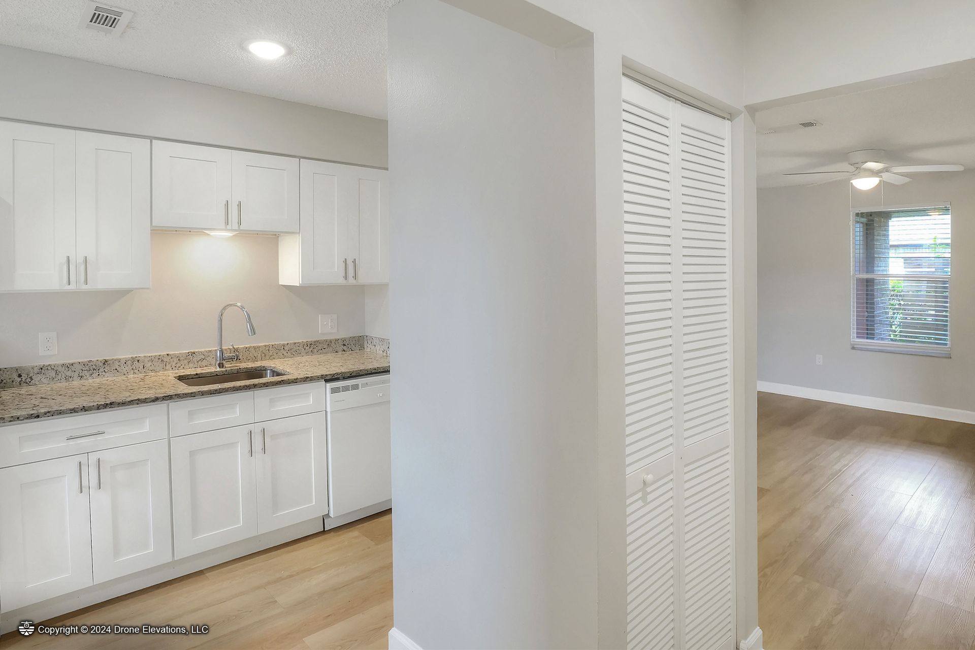 Kitchen with white cabinets, granite counters, next to a hall with a white closet and a living room with a window.