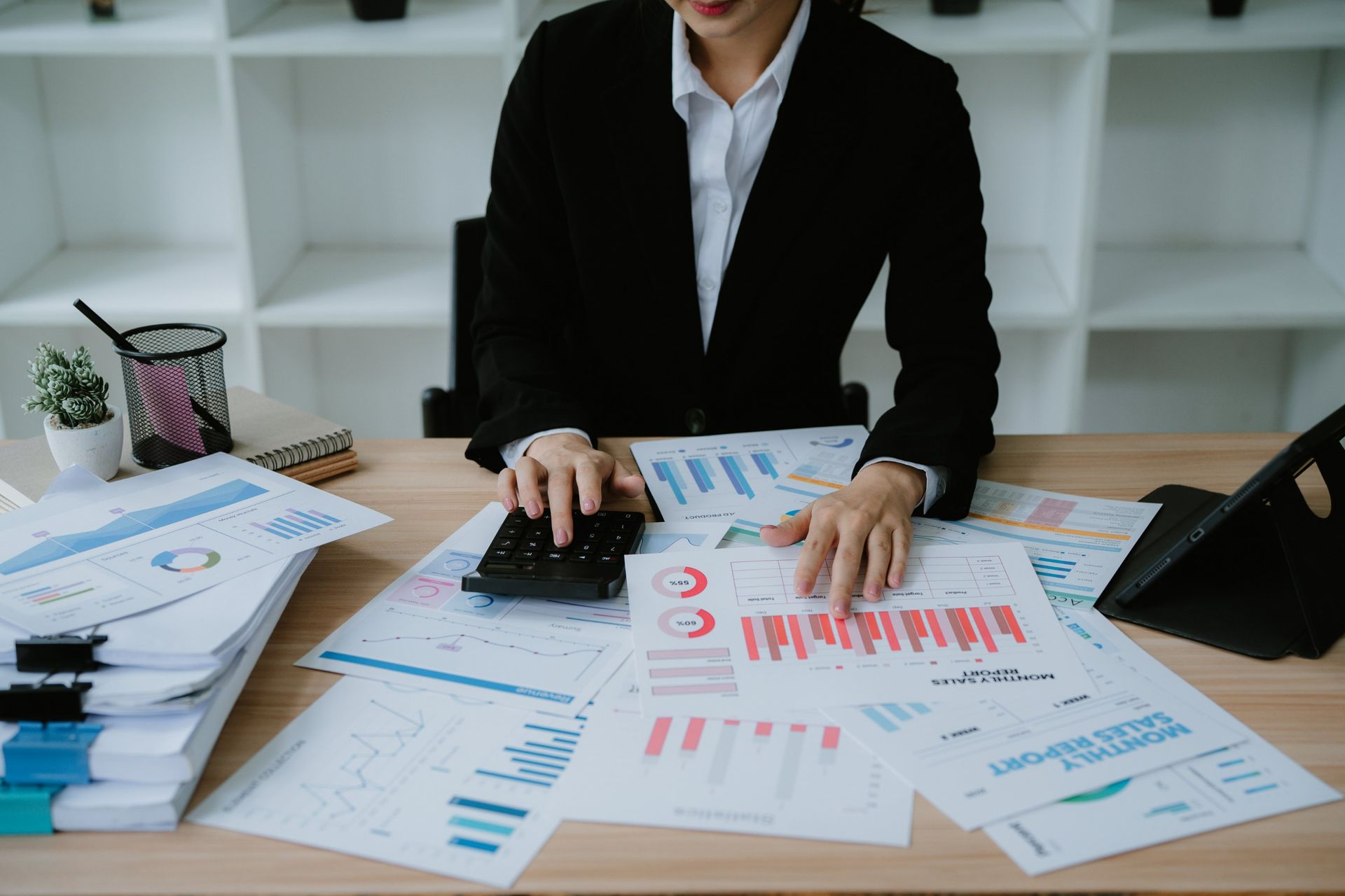 Person in a blazer using a calculator, surrounded by charts and financial reports on a desk.
