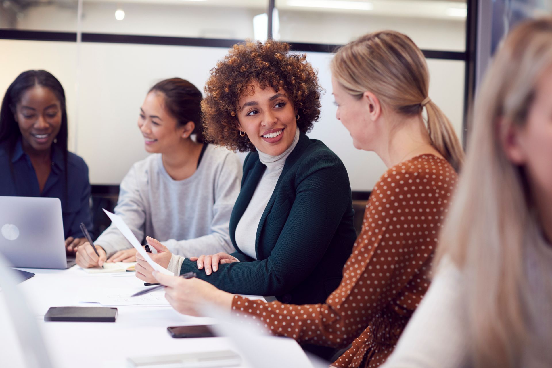 Four women in a meeting, smiling and discussing documents around a table. Laptop visible.