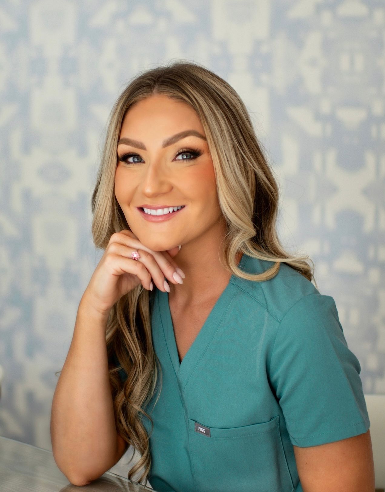A woman in a green scrub top is smiling with her hand on her chin.