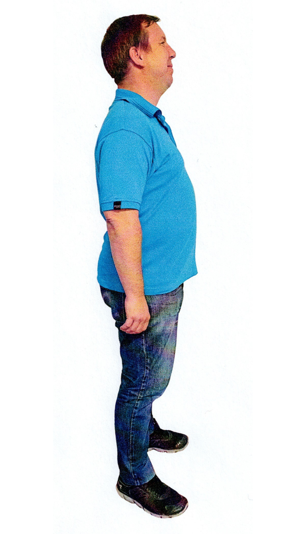 Lächelnde Frau, trägt ein schwarzes Hemd, einen Schal und blaue Jeans, die Hände in den Taschen, steht vor weißem Hintergrund.