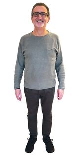 Mann mit Brille, grauem Langarmhemd und dunkler Hose, steht lächelnd vor weissem Hintergrund.