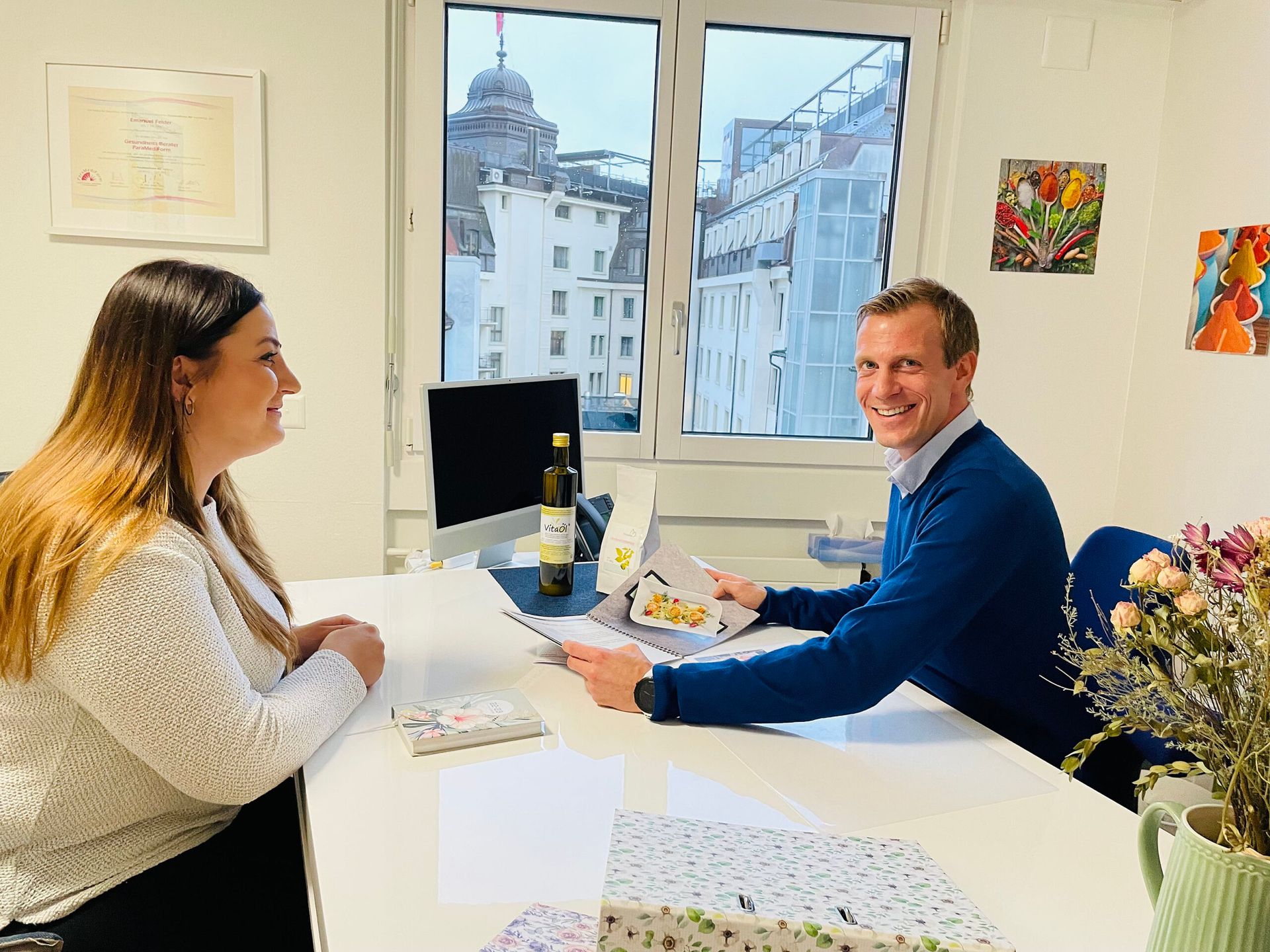 Ein lächelnder Mann im blauen Pullover sitzt einer Frau an einem Schreibtisch in einem Büro gegenüber.