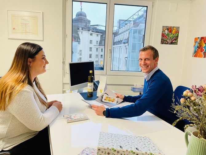 Ein lächelnder Mann im blauen Pullover sitzt einer Frau an einem Schreibtisch in einem Büro gegenüber.