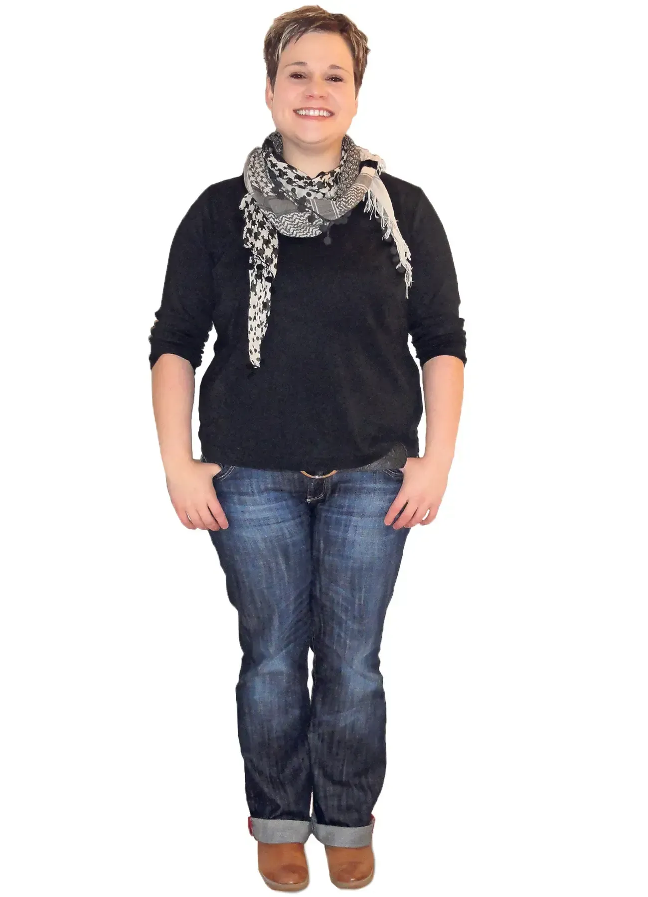 Lächelnde Frau, trägt ein schwarzes Hemd, einen Schal und blaue Jeans, die Hände in den Taschen, steht vor weißem Hintergrund.