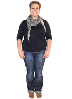 Lächelnde Frau, trägt ein schwarzes Hemd, einen Schal und blaue Jeans, die Hände in den Taschen, steht vor weißem Hintergrund.