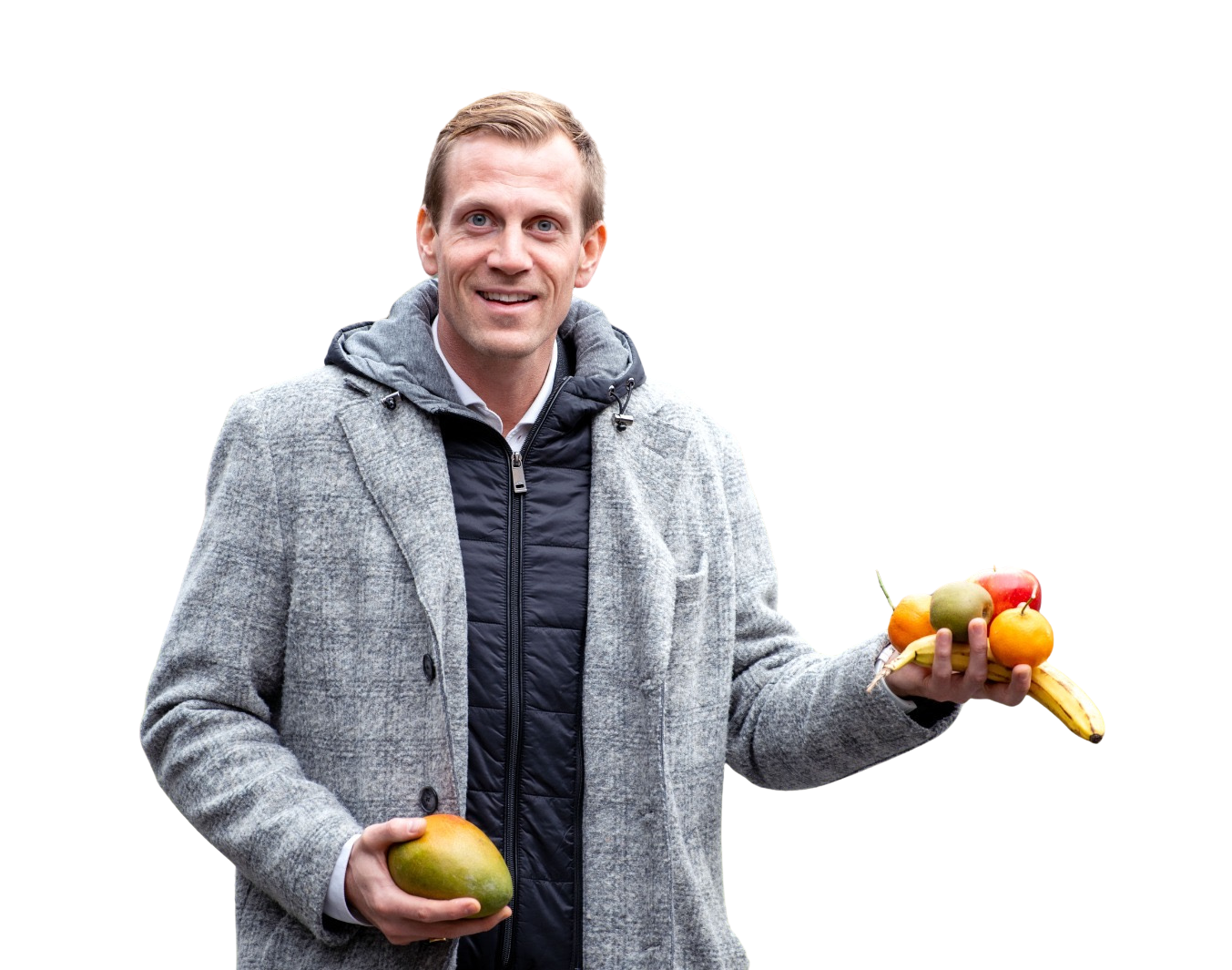 Ein Mann hält eine Mango und verschiedene Früchte in der Hand; er lächelt. Er befindet sich im Freien und trägt einen grauen Mantel und eine schwarze Weste.