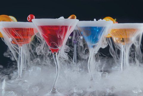 Une rangée de verres à martini remplis de liquides de différentes couleurs et de fumée qui en sort.