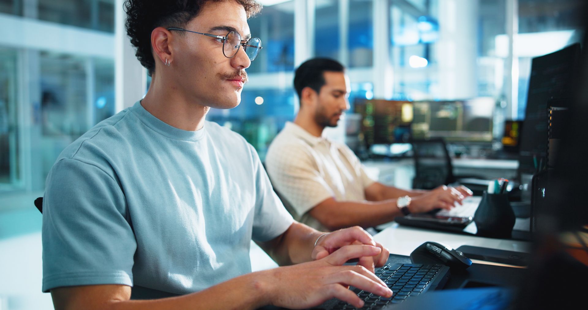 Two men working on computers in a modern office setting. One typing, wearing glasses.