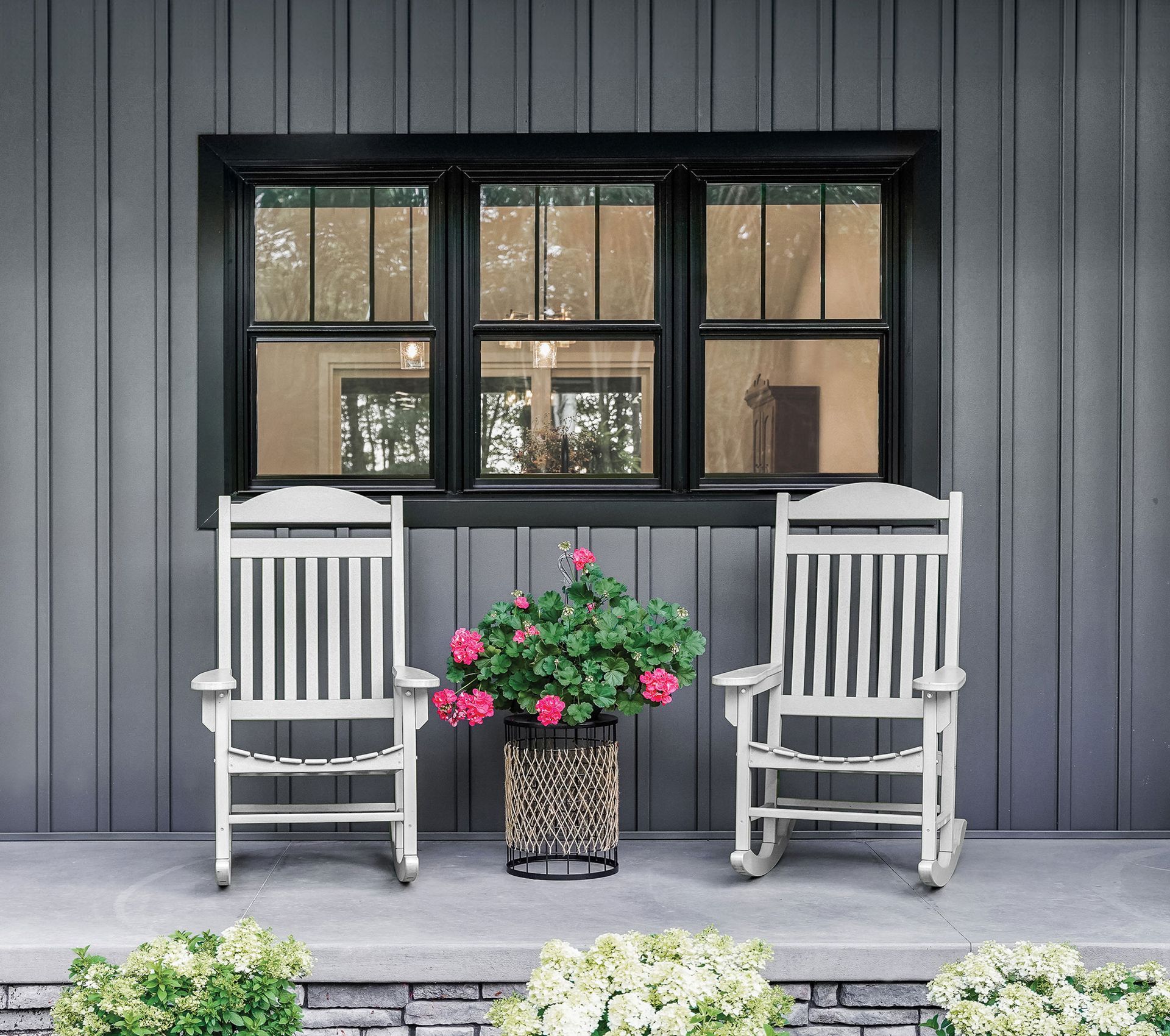 Two white rocking chairs on a porch with a flower basket, set against a gray-sided wall with a black-framed window.