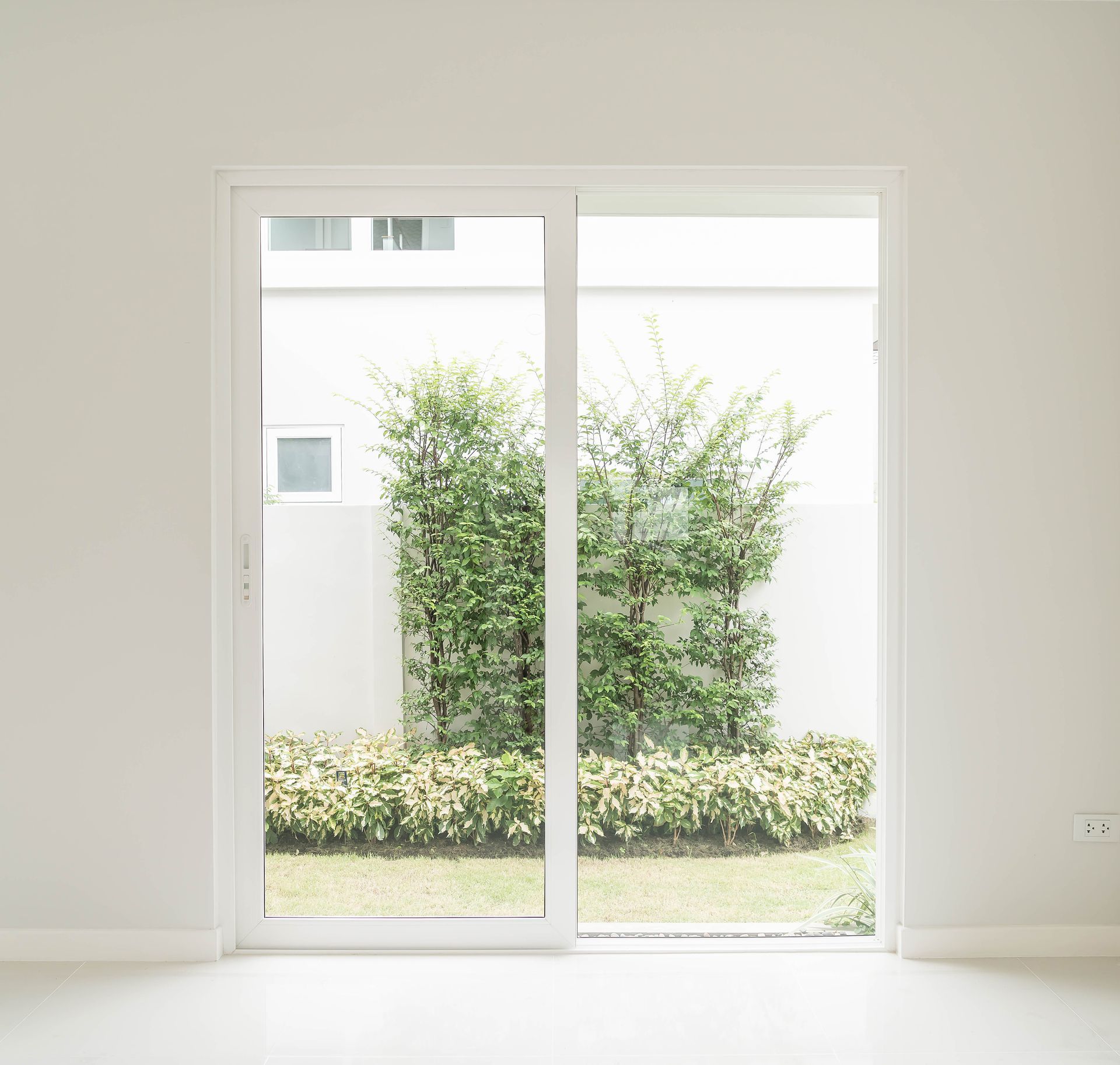 White sliding glass door leading to a yard with green plants against a white wall.