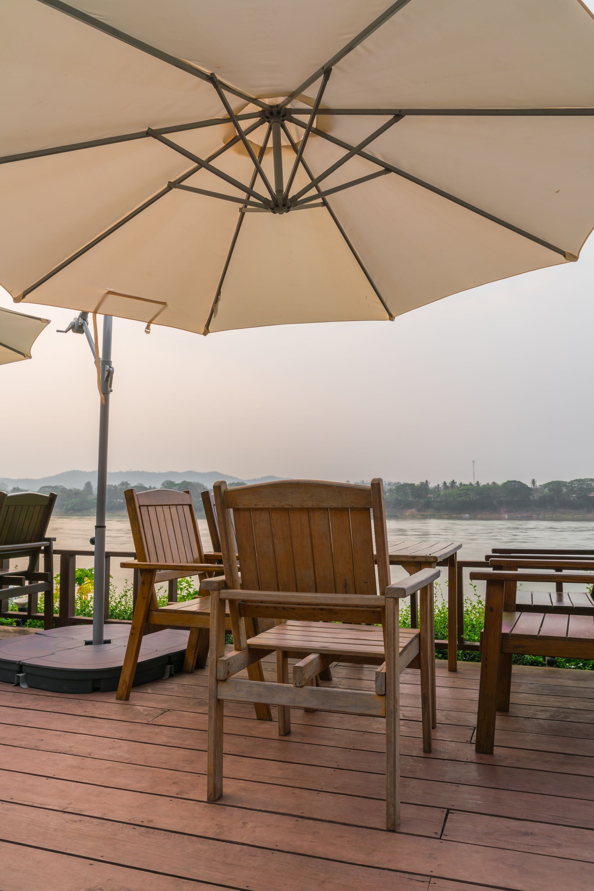 Outdoor cafe seating area with umbrella overlooking water. Wooden chairs, tables, and a tan umbrella provide shade.