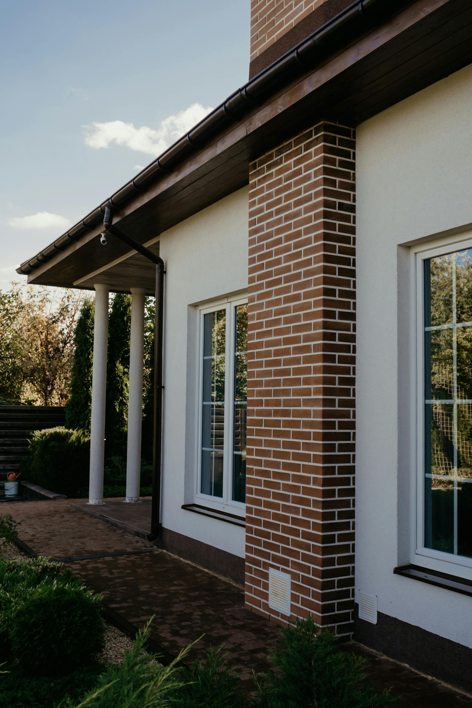 Exterior of a white house with a brick column, white-framed windows, and a dark brown roof.