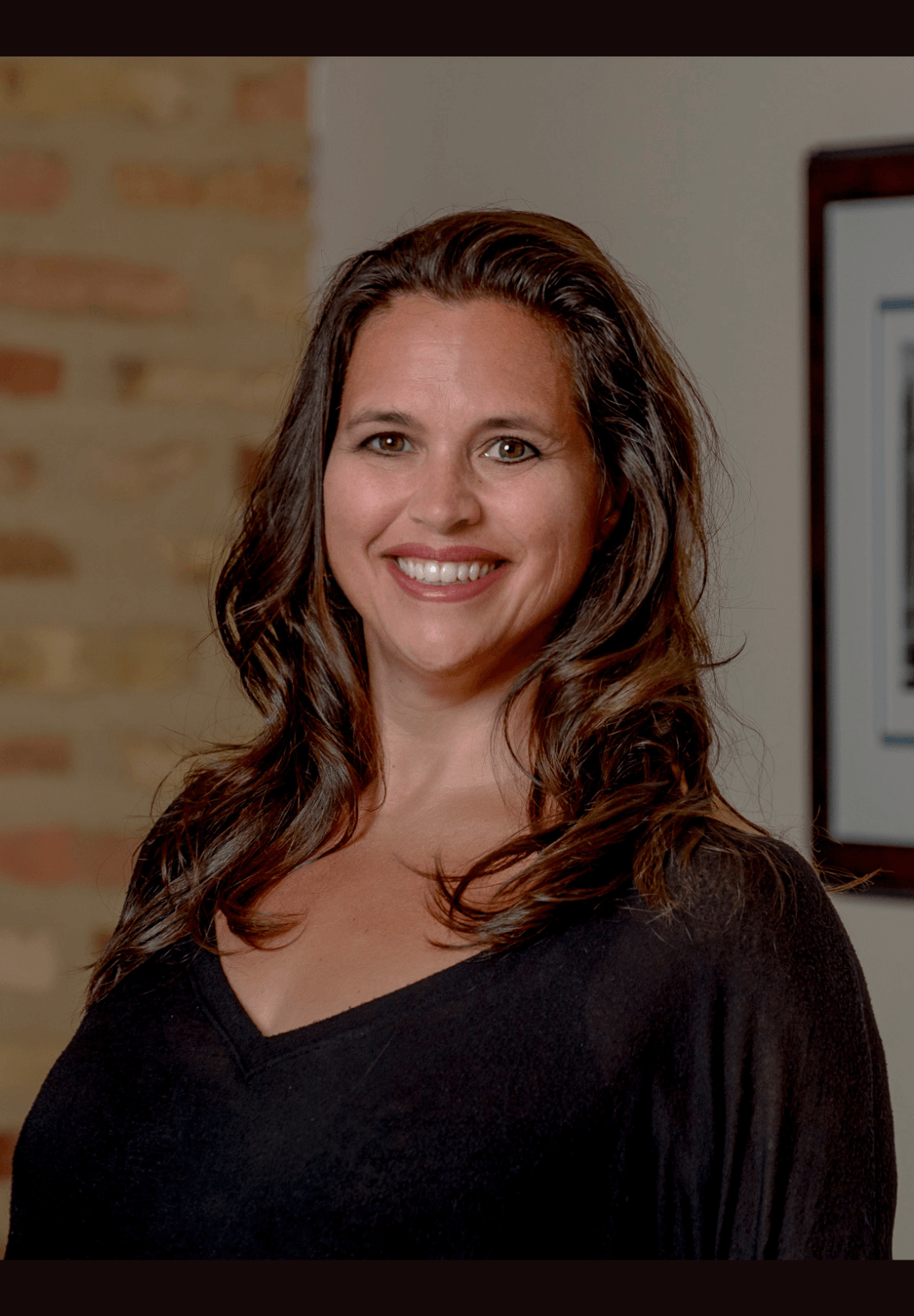 A woman in a black shirt is smiling in front of a brick wall.