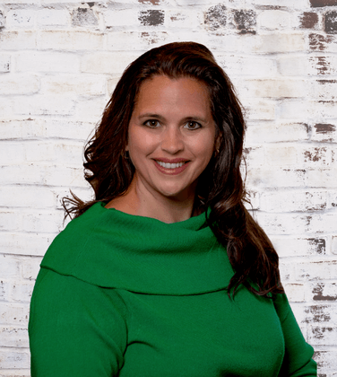 A woman in a green sweater is smiling in front of a white brick wall.