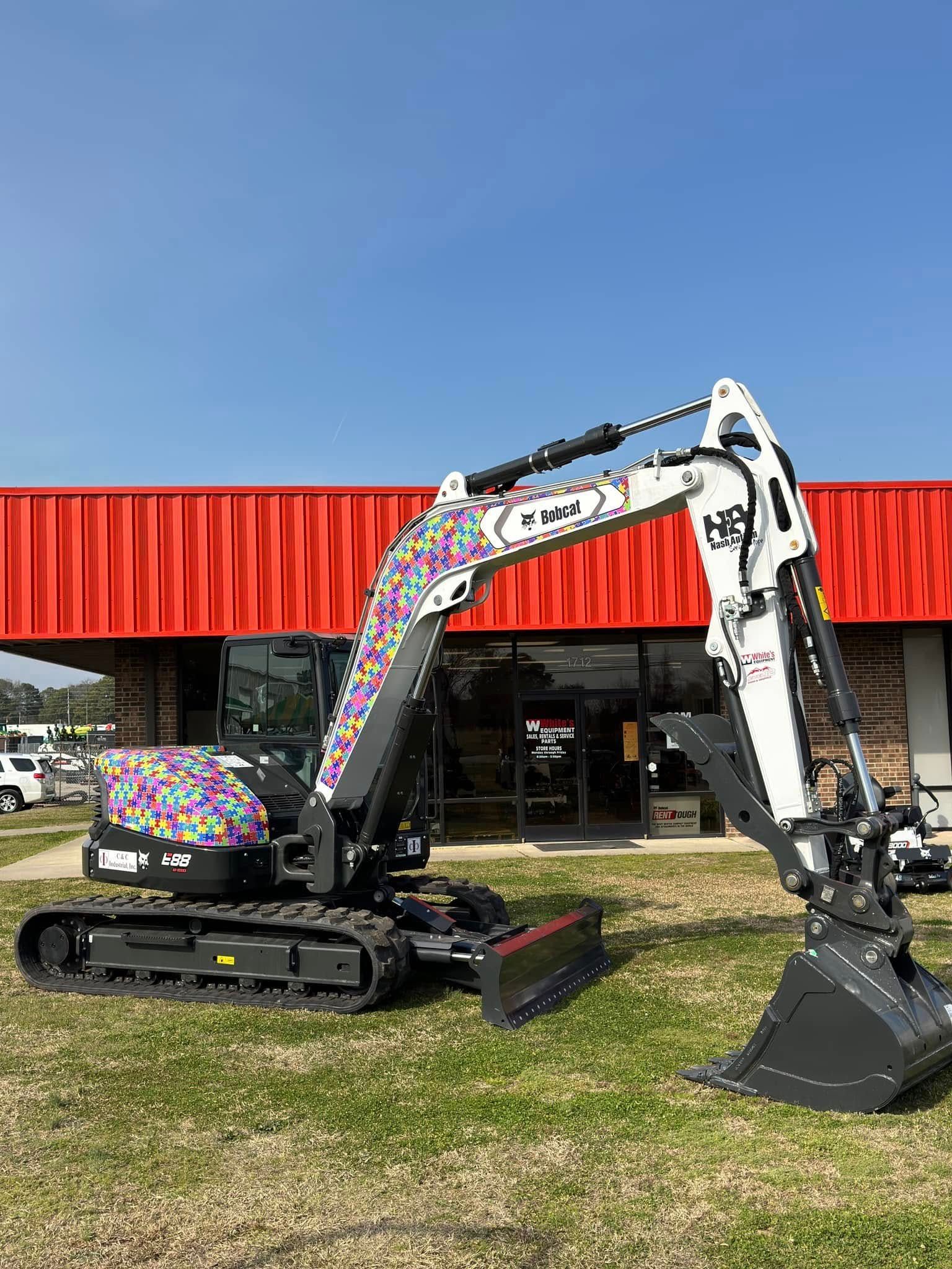Bobcat excavator with colorful accents parked on grass in front of a building with a red facade.