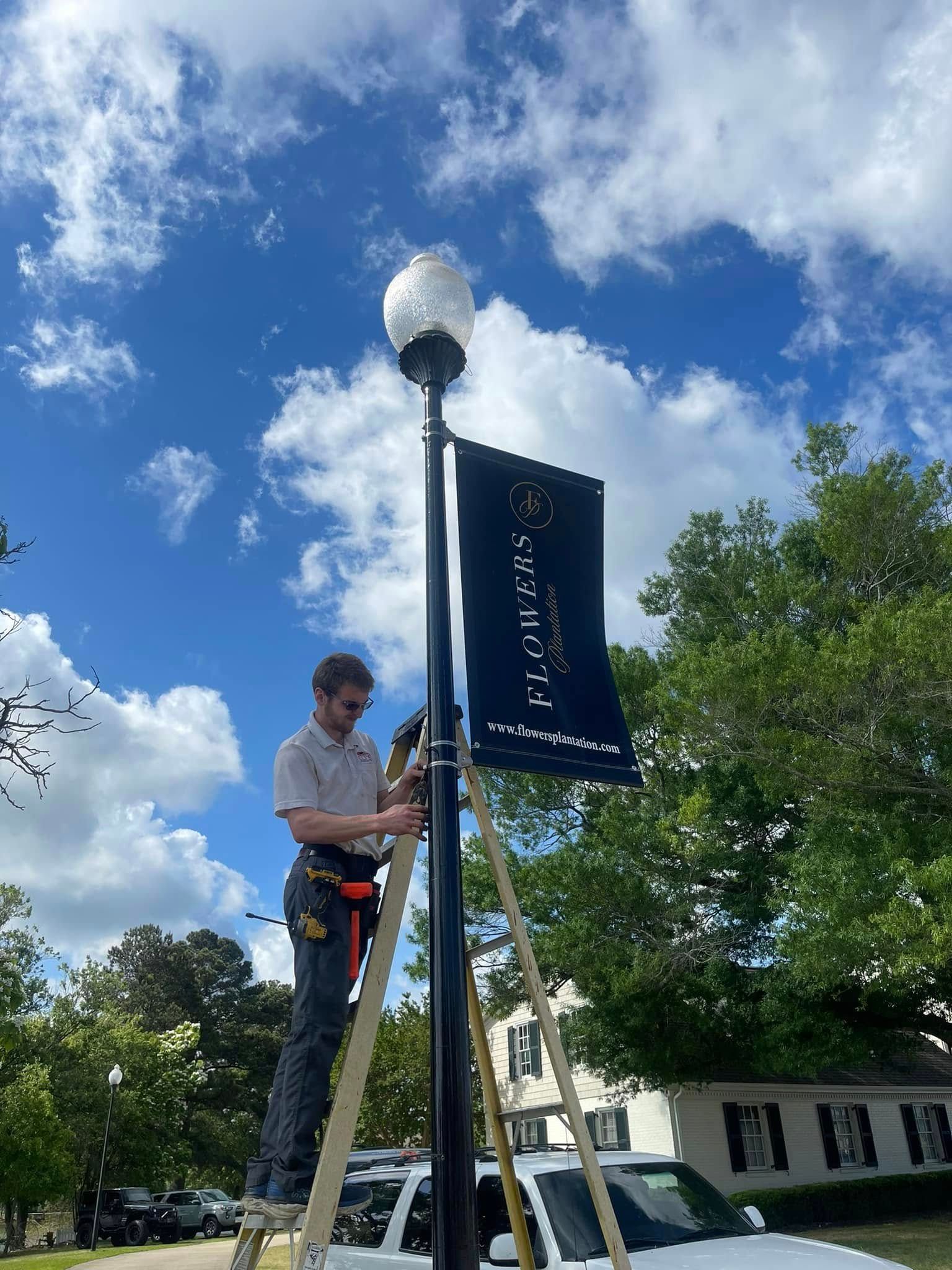 A man on a ladder installs a banner on a black lamppost under a blue sky.