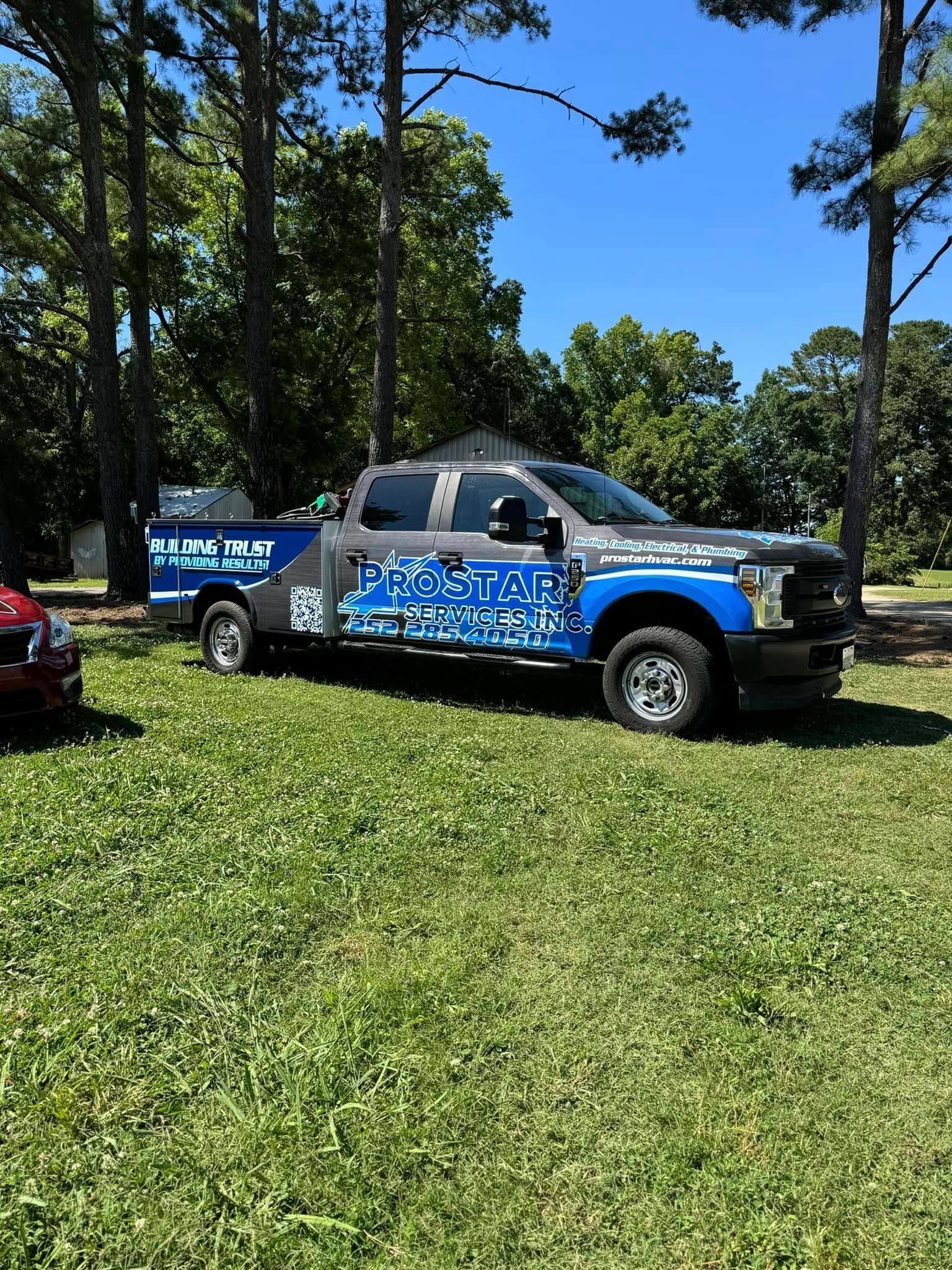 Blue and grey wrapped truck parked on grass, blue sky and trees in background.