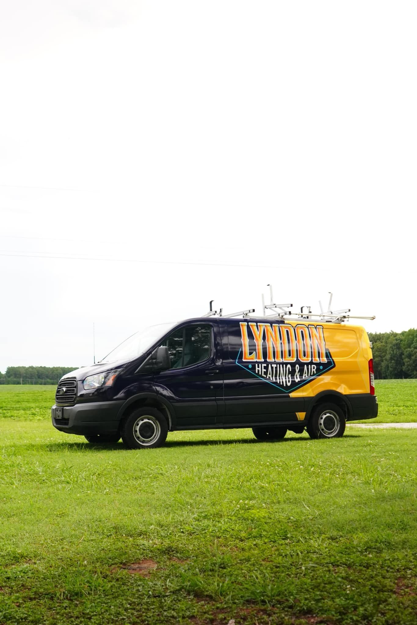 Dark blue and yellow work van parked on green grass field. Lettering on the side. Overcast sky.