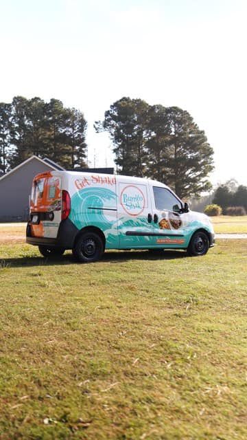 A white food truck with turquoise and orange graphics is parked on grass in front of a building.