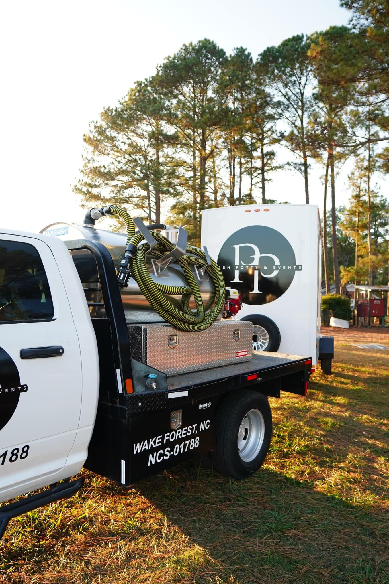 White truck with trailer in a grassy area, trees in the background, logo on the truck.