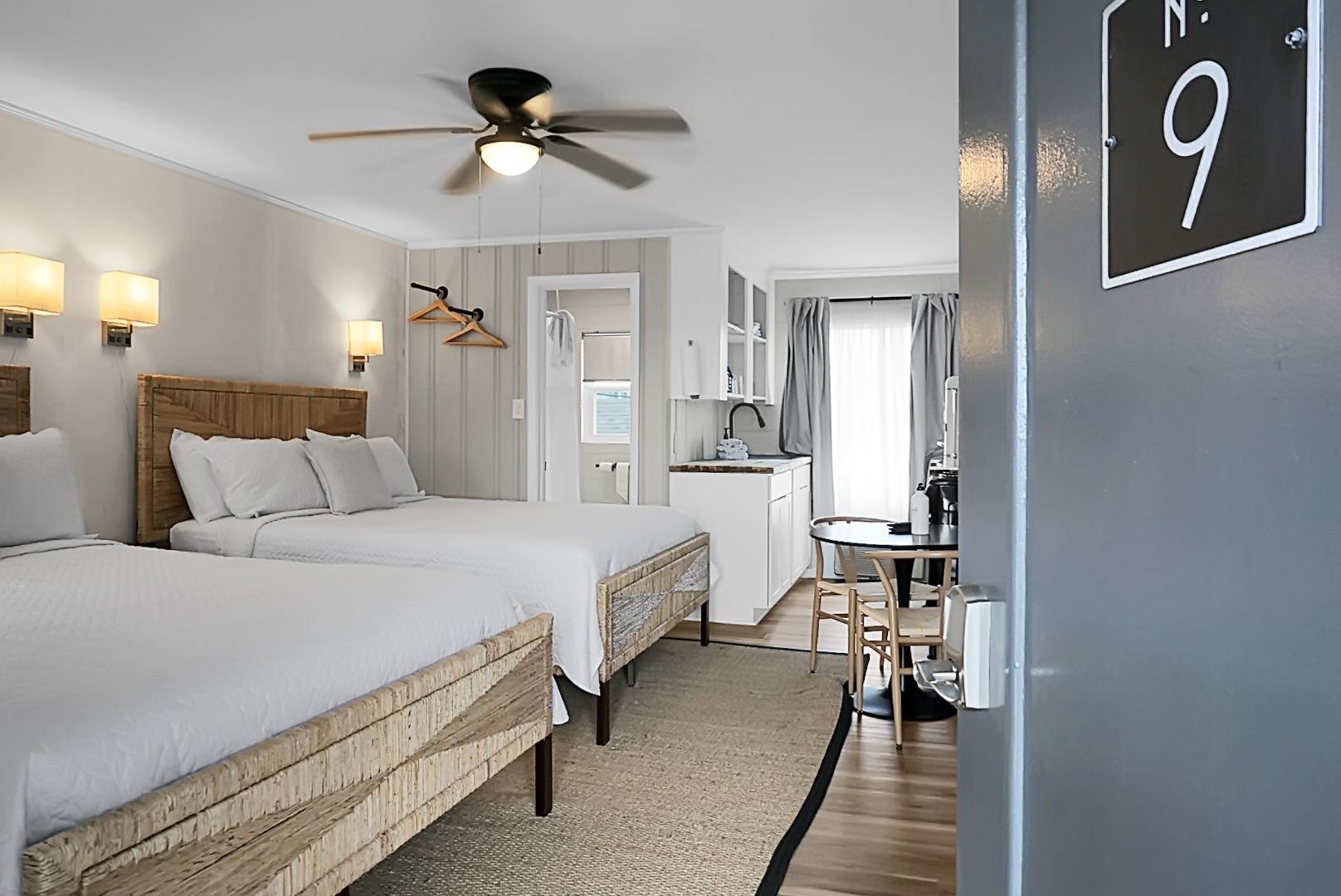 A bright and airy guest room at Seabirds Motel at Kure Beach featuring a spacious bed with white linens, a woven rattan bed frame, a ceiling fan, and light-colored furnishings. The room offers a clean, coastal-inspired ambiance, perfect for a pet-friendly stay in North Carolina.